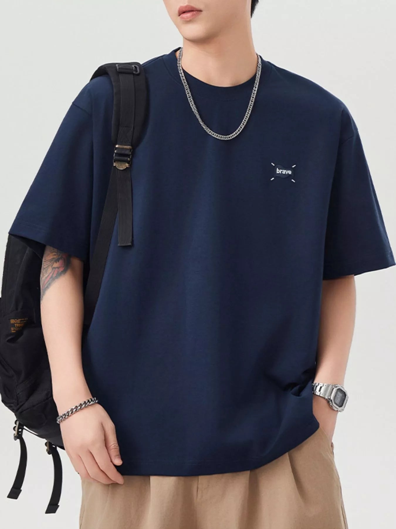 Person wearing a navy blue t-shirt with a small logo, beige pants, and a black backpack on a light gray background.