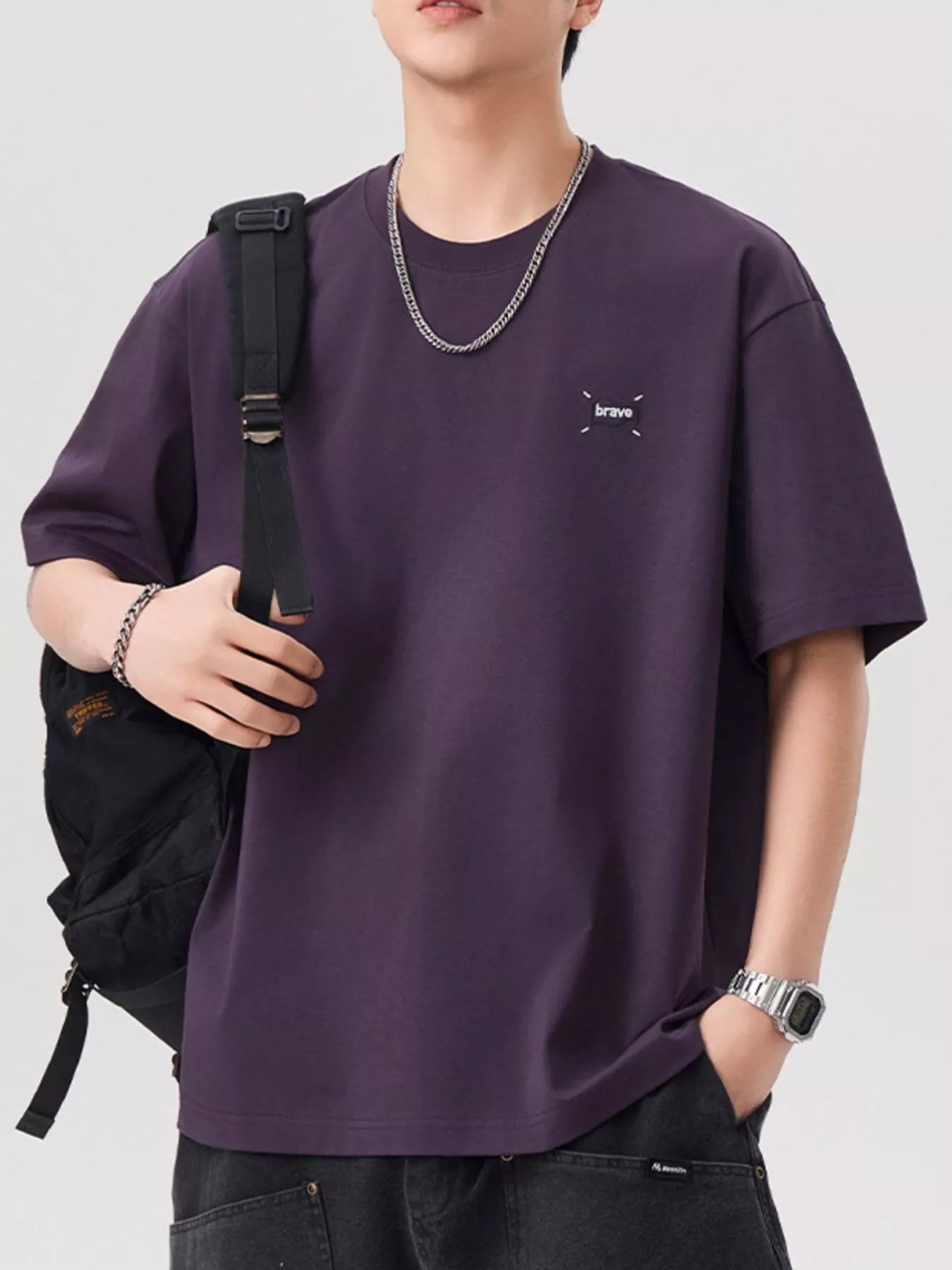 Person wearing a purple t-shirt with a small logo, holding a black backpack on a white background